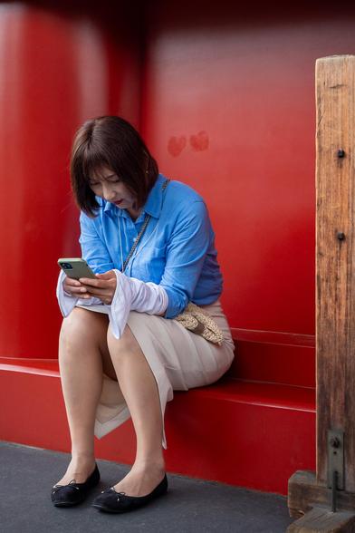 A young woman with black hair, all made up and wearing a light beige skirt and a light blue shirt with sleeves, texting on her mobile phone, while seating on a step with dark red painted wall in the background. Above her head, visible traces of two heart shapes which seem to have been removed from the wall and painted in the same color in an attempt to hide them.