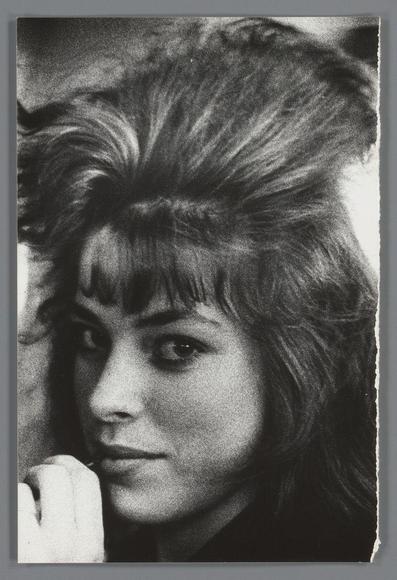 The image is a black and white portrait of an individual with striking, voluminous hair styled in multiple directions. The hairstyle has been described as reminiscent of the "beehive" or punk era fashion popularized in the 1970s. The person's facial features are partially visible; they have intense eyes looking directly at the camera, and a hint of their cheek is seen beneath an arm that appears to be touching their face. There is no discernible background detail as it seems cropped tightly around the subject, emphasizing the hairstyle and expression over any contextual information. This style of hair was associated with avant-garde fashion trends during its time.