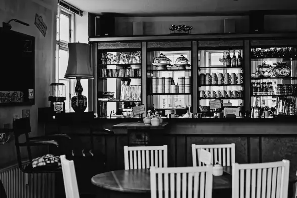 Ein Blick in ein leeres Kaffee. Tassen und Teller und was man so alles hinter dem Tresen braucht, stehen ordentlich, sauber und in Reih und Glied in den Regalen. Am Tisch im Vordergrund stehen vier Stühle. Von links fällt durch ein Fenster das Tageslicht herein, sodass der Vordergrund recht dunkel wirkt, der Hintergrund mit den Regalen jedoch hell erleuchtet.