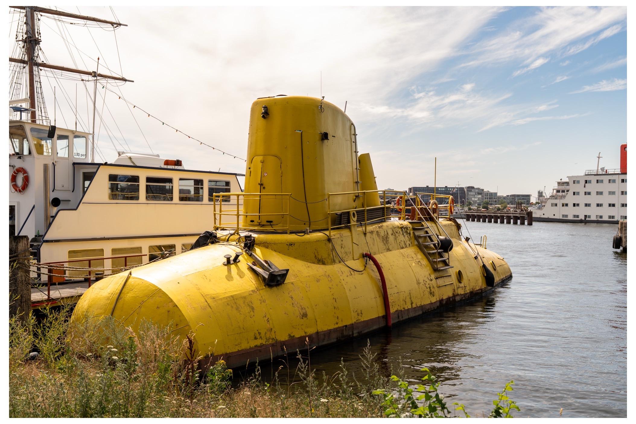 A large yellow submarine is docked near other boats in a harbor. The scene features clear skies and calm waters, with greenery visible in the foreground.
