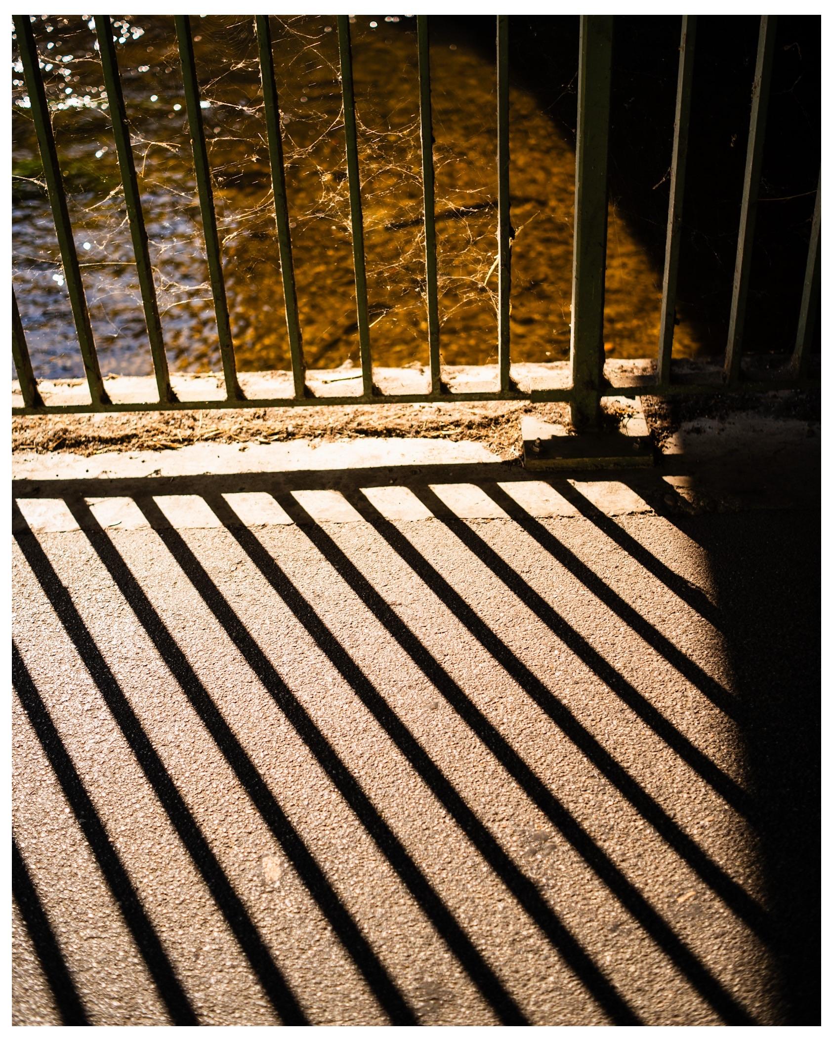Ein Geländer durch das die Sonne scheint samt seiner Schatten auf dem Asphalt.