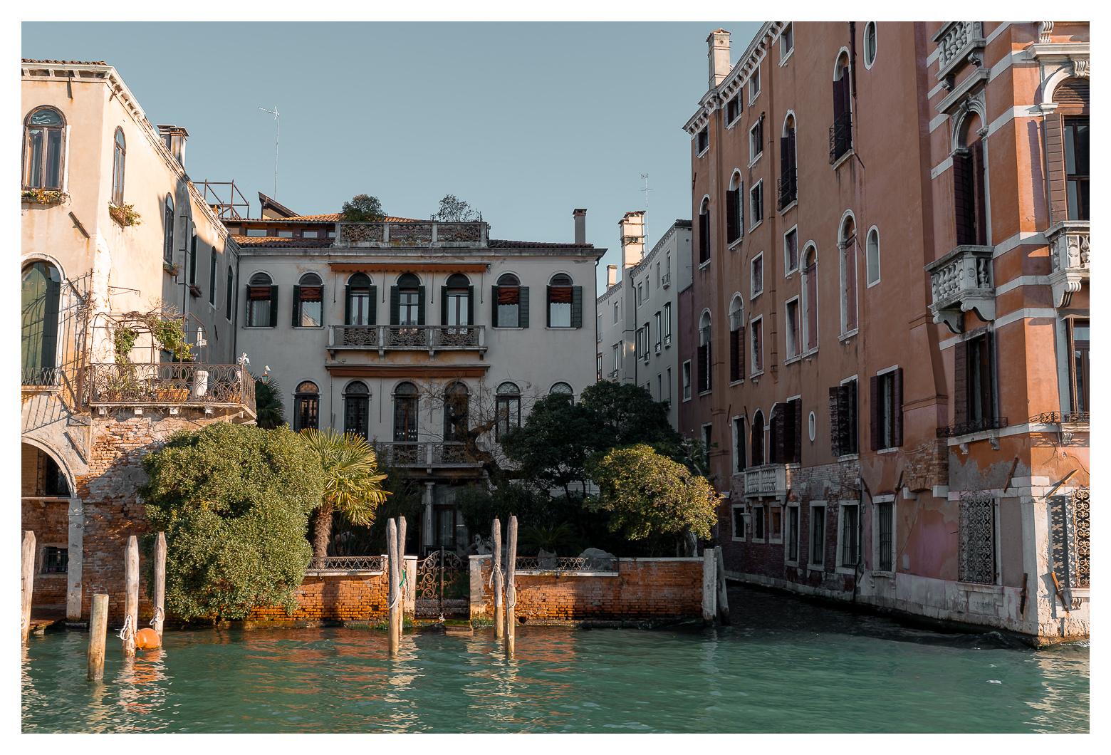 Szene aus Venedig vom Wasser aus aufgenommen: ein kleiner ummauerter Garten am Kanal, dahinter ein Palazzo. Links und rechts weitere Pallazi und ein schmaler Kanal links. Gedeckte Farben, viel Orange/Rot was einen kontrast mit dem Wasser bildet.