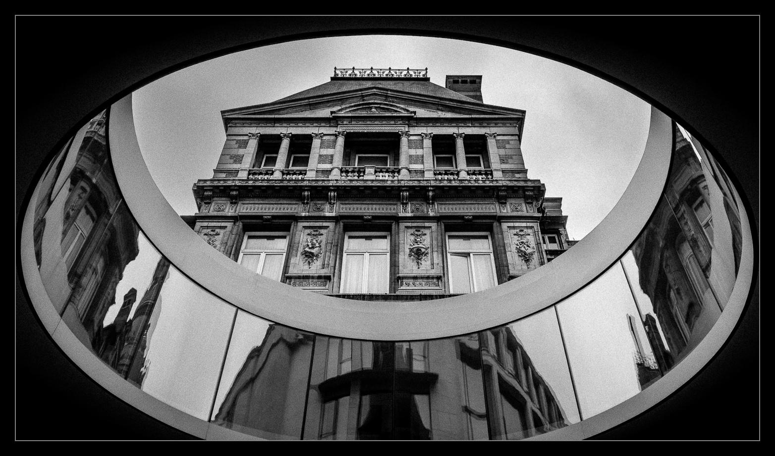 Blick von unten nach oben auf ein historisches Gebäude. Die besonderheit ist, dass zwischen diesem und dem Gebäude im Rücken des Fotografen eine Art Brücke/Verbindung in Form eines Rings geschaffen wurde. Wir blicken durch den Ring hoch zum Gebäude.