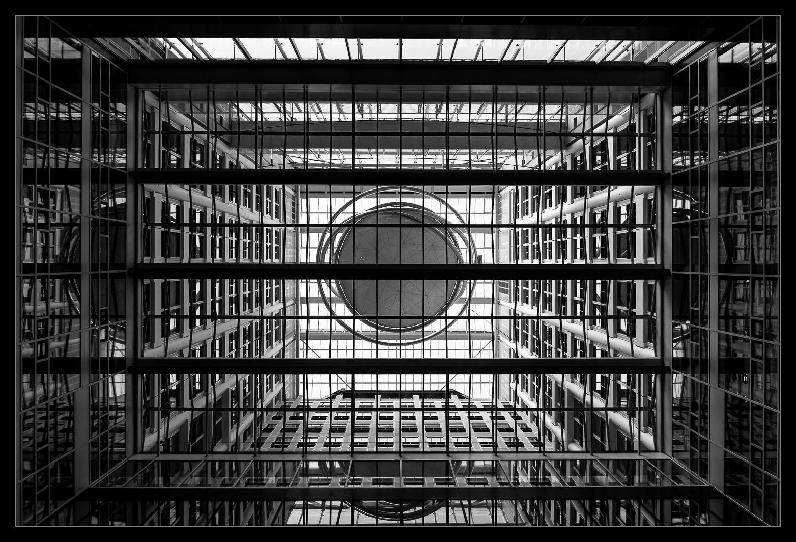 Blick Senkrecht nach oben in einem großen modernen Gebäude. Wir sehen viel Glas, ein mitten im Raum schwebendes Kunstwerk und so etwas wie Verbindungsbrücken zwischen Gebäudeteilen.