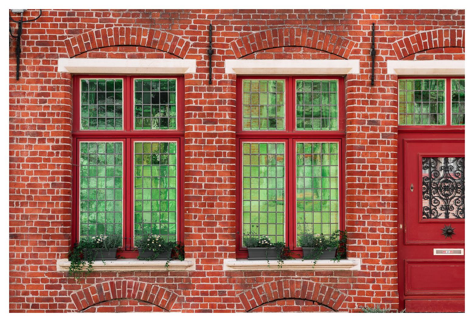 Blick auf ein Klinkerhaus mit roter Tür und rot gerahmten Bleiglasfenstern. Die scheiben haben einen eigentümlichen grün-schillernden Farbton, der mit dem restlichen rot sehr schön kontrastiert.