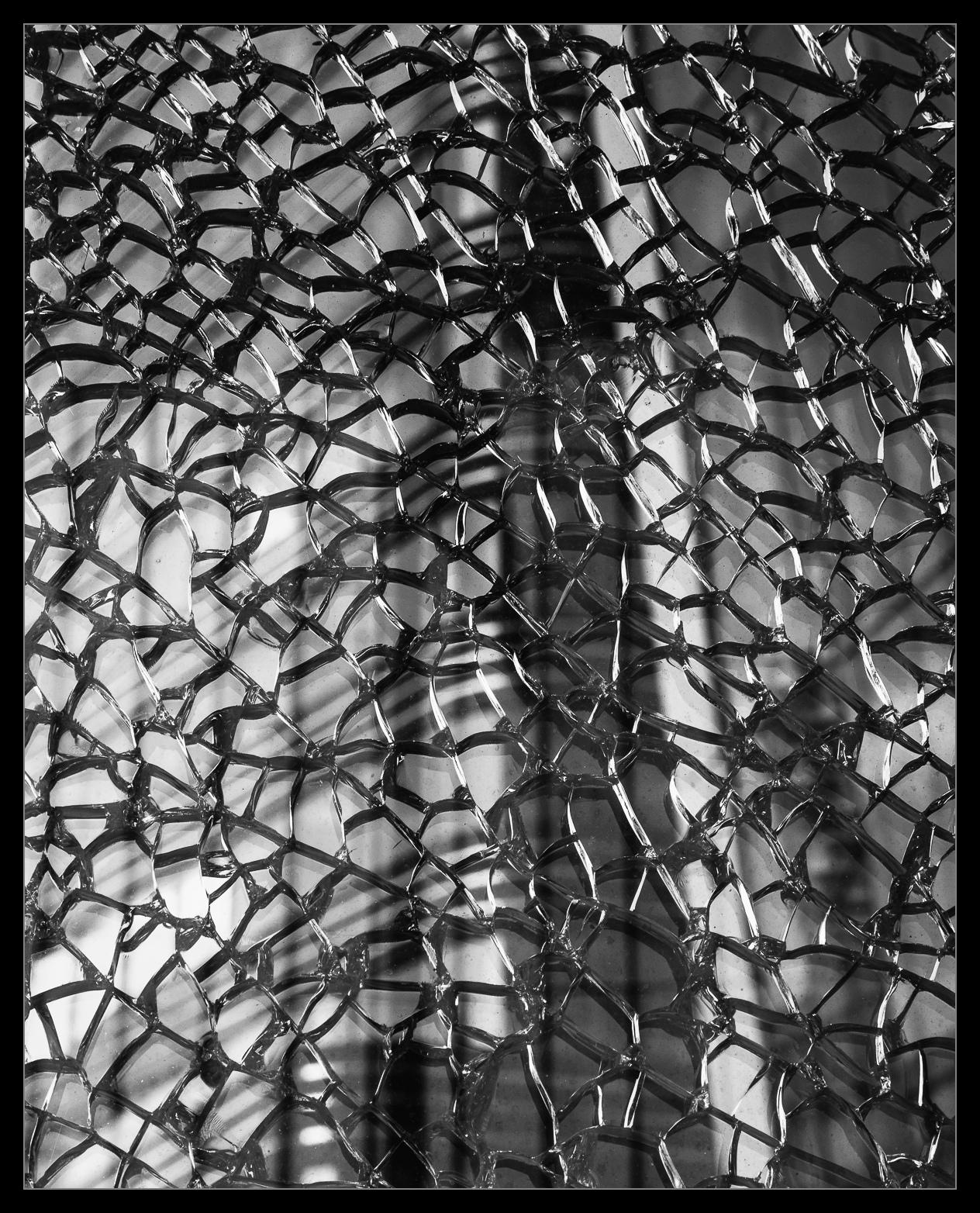 Zerbrochene Glasscheibe im Fokus, dahinter im Bokeh (schwer zu erkennen) ein Gebäude mit Glasfront.
